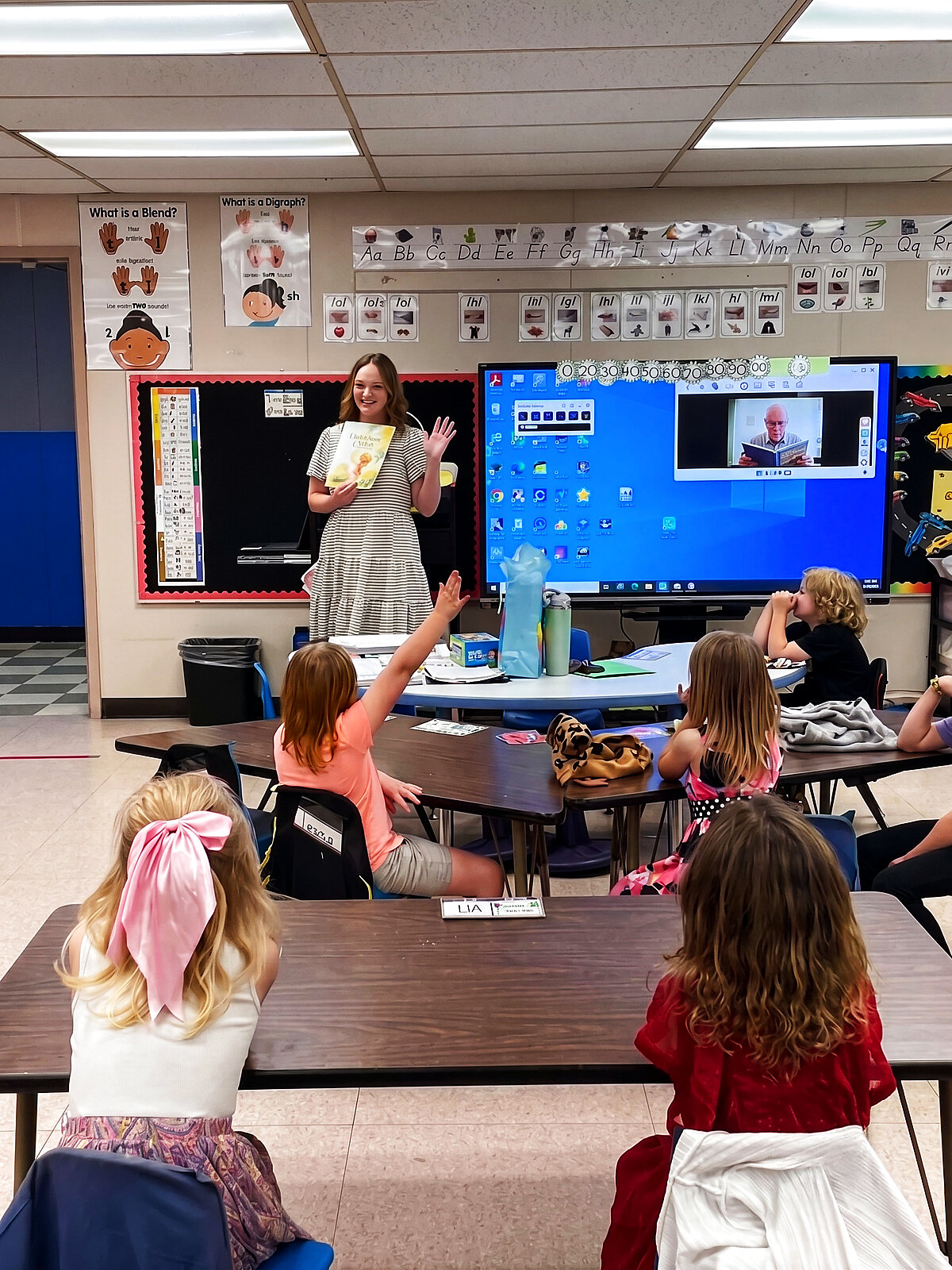 Brandi Filer holding up a book while reading aloud to a classroom, with a guest reader joining by video call on the smartboard